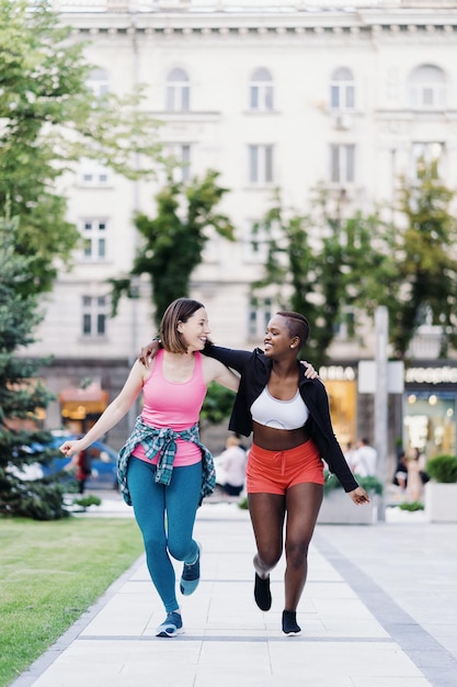 Fröhlich lächelnde Freunde in Sportbekleidung, die in der Stadt laufen und multiethnische Frauen beim Fitnesstraining diskutieren