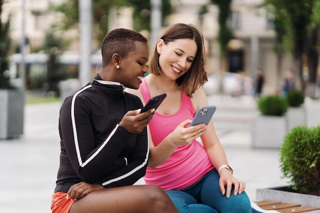 Fröhlich lächelnde Freunde in Sportkleidung, die auf einer Bank in der Stadt sitzen und diskutieren, während sie mit dem Smartphone versuchen, multiethnische Frauen zu screenen, die eine Fitness-Workout-Pause haben