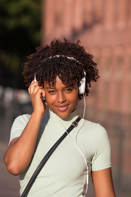 Fröhlich lächelnde lateinamerikanische Frau mit lockigem Haar, die Musik auf dem Smartphone hört, trägt Kopfhörer auf der Straße und blickt in die Kamera