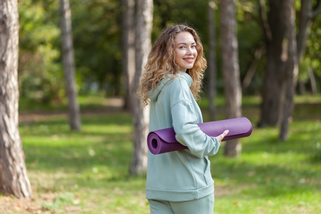 Fröhlich lächelnde Passform einer kaukasischen Frau mit lockigem Haar, die an einem sonnigen Tag im Park eine Yogamatte in der Hand hält Gesunder Lebensstil Outdoor-Training