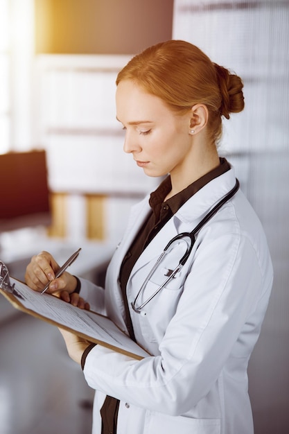 Fröhlich lächelnde Ärztin mit Klemmbrett in sonniger Klinik. Porträt einer freundlichen Ärztin bei der Arbeit. Medizin-Konzept.