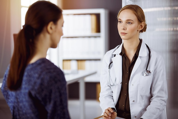 Fröhlich lächelnde Ärztin und Patientin diskutieren aktuelle Gesundheitsuntersuchungen, während sie in einer sonnigen Klinik sitzen. Medizin-Konzept.