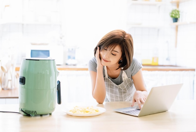 Fröhlich lächelnde schöne asiatische Frau, die an einem Lalaptop-Computer arbeitet, während sie zu Hause mit Luftfritteusen in der Küche kocht, während sie sich selbst von der Ausbruchskrise des Coronavirus isoliert Neues normales Lebensstilkonzept