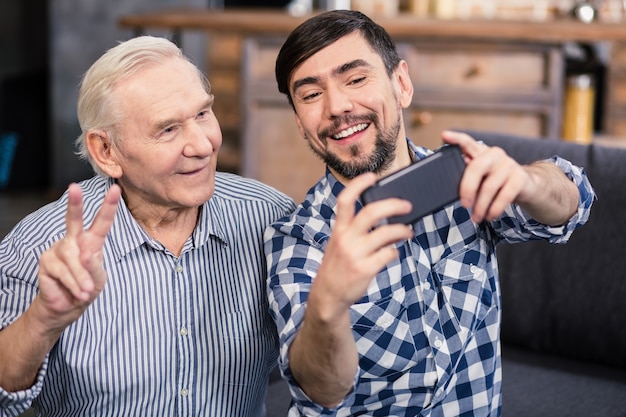 Fröhlich lächelnder alter Mann, der vor der Kamera posiert, während er Selfies mit seinem Sohn macht