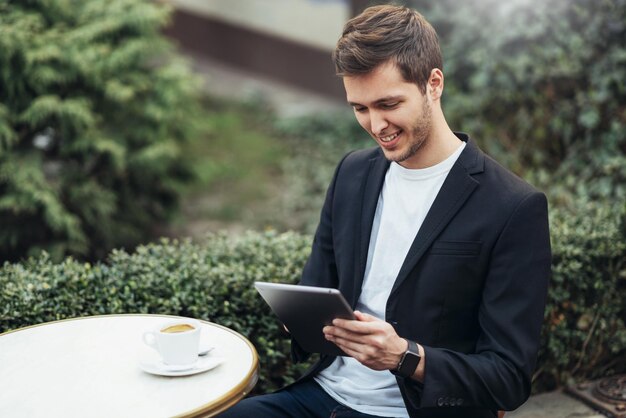 Fröhlich lächelnder kaukasischer Student mit Tablet-Gerät, der im Internet surft und Newsfeeds in sozialen Medien während der Kaffeepause im modernen Café in der Stadt überprüft Technologie- und Geschäftskonzept