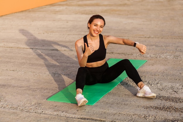 Fröhlich lächelndes athletisches Mädchen, das den Fitness-Tracker der Armbanduhr zur Hand zeigt und mit den Daumen nach oben gestikuliert, zufrieden mit den Gesundheitsindikatoren nach dem Training auf der Yogamatte Sportaktivität im Freien