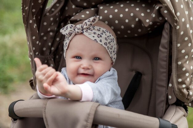 Fröhlich lächelndes emotionales acht Monate altes blauäugiges Mädchen sitzt im Kinderwagen auf einem Spaziergang und wartet auf Mama. Kleines und süßes Baby mit Stirnband auf dem Kopf, das im Kinderwagen sitzt. Konzept der richtigen Erziehung und Kindheit