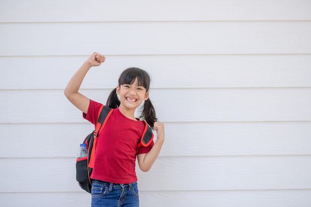 Fröhlich lächelndes kleines Mädchen mit großem Rucksack springt und hat Spaß gegen die weiße Wand. Blick in die Kamera. Schulkonzept. Zurück zur Schule