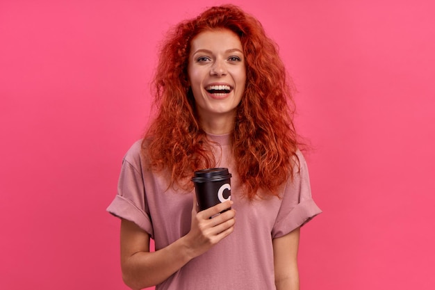 Fröhlich lächelndes rothaariges hübsches Mädchen hält Kaffee zum Mitnehmen auf rosa Hintergrund