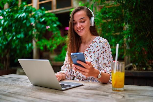 Fröhlich lächelndes schönes süßes junges tausendjähriges Freiberuflermädchen, das drahtlose Kopfhörer mit Laptop und Telefon für Remote-Arbeit, soziale Netzwerke und Online-Browsing im grünen Café trägt