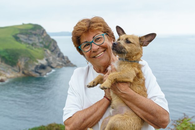 Fröhliche ältere Frau, die einen braunen Hund hält. Horizontale Ansicht der älteren Frau mit ihrem Hund an Feiertagen im Freien. Ruhestandsleute und Hundekonzept.