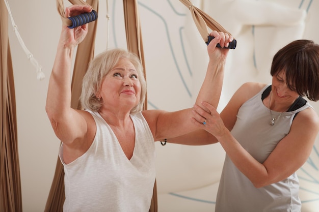 Fröhliche ältere Frau lächelt, während sie Aerial Yoga auf der Hängematte mit Personal Trainer praktiziert