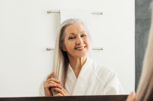 Fröhliche ältere Frau mit grauen Haaren, die sich im Badezimmer einen Spiegel ansieht