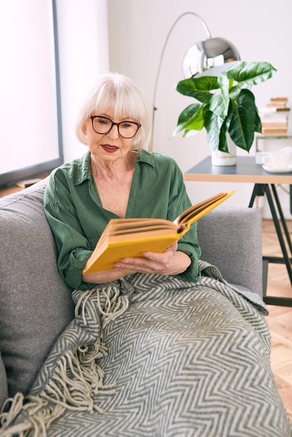 Fröhliche ältere Frau sitzt auf der Couch und liest zu Hause ein Buch