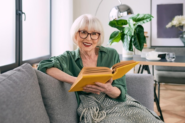 Fröhliche ältere Frau sitzt auf der Couch und liest zu Hause ein Buch