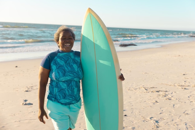 Fröhliche afrikanisch-amerikanische ältere Frau mit Surfbrett, die am Strand gegen Meer und klaren Himmel steht.