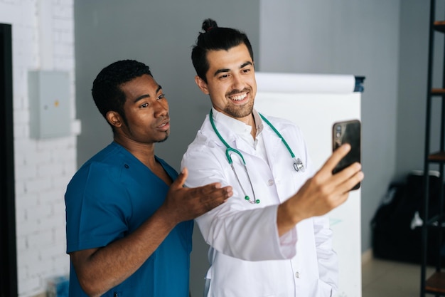 Fröhliche afrikanische und kaukasische Männer in medizinischer Uniform, die ein Selfie auf dem Smartphone machen und im Konferenzbüro des Krankenhauses stehen. Zwei glückliche medizinische Mitarbeiter, die nach einem internationalen Treffen ein Foto machen.