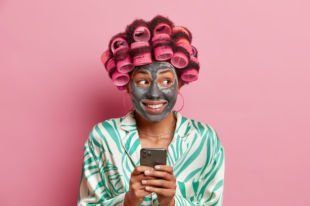 Fröhliche Afro-Amerikanerin lächelt positiv wendet Tonmaske an verwendet Handy zum Surfen im Internet macht perfekte Frisurgekleidet in häuslicher Robe isoliert über rosa Wand