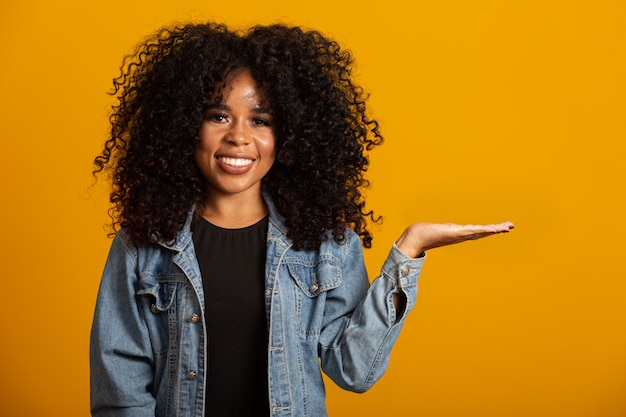 Fröhliche Afro-Frau zeigt auf Kopierfläche, bespricht erstaunliche Promo, gibt nach oder Richtung, trägt gelben warmen Pullover, hat ein angenehmes Lächeln, fühlt sich optimistisch, isoliert über gelber Wand.