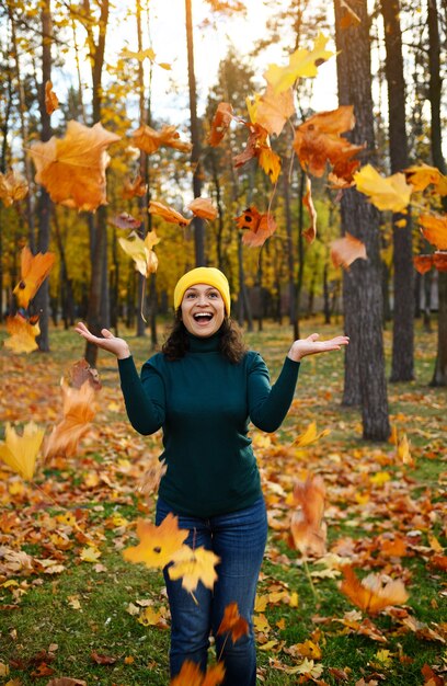 Fröhliche Afroamerikanerin, die einen warmen, gemütlichen, bunten, gelben Wollhut, einen grünen Pullover, einen lässigen Denim trägt, wirft gelb-orange, trocken gefallene Ahornblätter im Herbst, genießt das Herbstwetter im schönen Park.