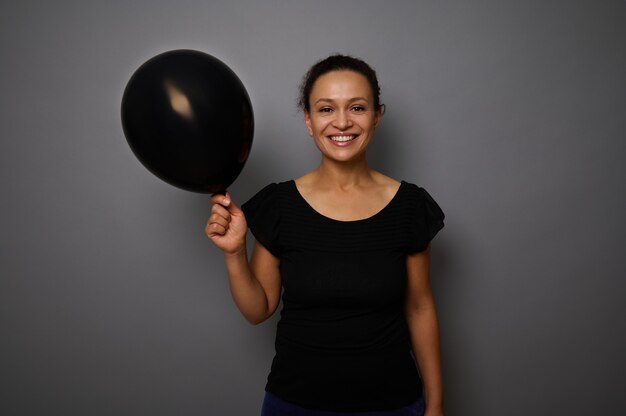 Fröhliche afroamerikanerin in schwarz gekleidet lächelt in die kamera und posiert vor grauem hintergrund mit einem schwarzen luftballon in der hand. black friday-konzept mit kopien-werbefläche