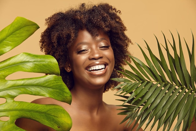 Fröhliche Afroamerikanerin mit gesunder, sauberer Haut, die gegen tropische Blätter steht