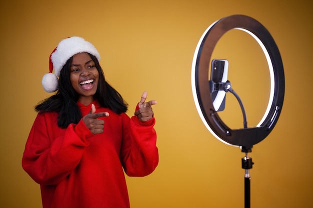 Fröhliche afroamerikanische Bloggerin in einem roten Hoodie und einer Weihnachtsmütze mit einer beleuchteten Ringlampe