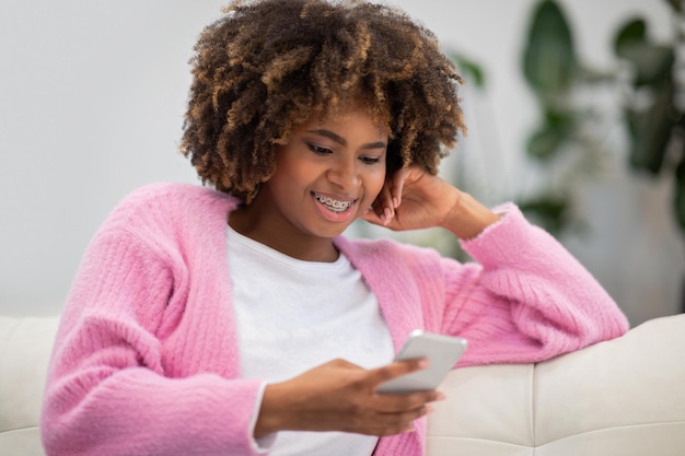 Fröhliche afroamerikanische Dame, die mit Smartphone auf der Couch sitzt