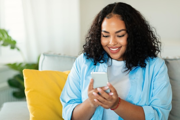 Fröhliche afroamerikanische Frau mit Smartphone zu Hause sitzen