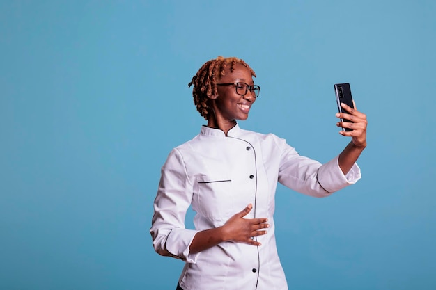 Fröhliche afroamerikanische Köchin bei einem Videoanruf mit Handy in Studioaufnahme. Professioneller Koch in Uniform, der auf einem Handheld-Gerät mit Freunden spricht, isoliert auf blau.