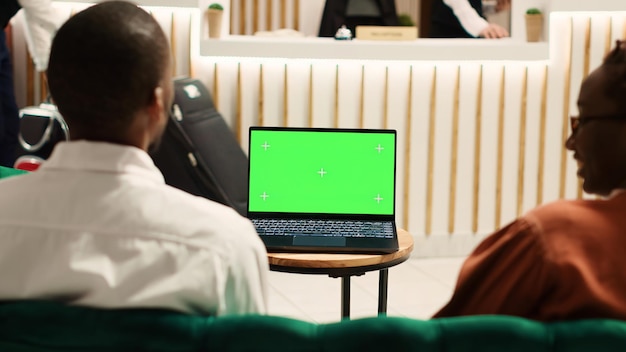 Fröhliche afroamerikanische Touristen, die sich im Hotelfoyer ein Video auf einem Laptop-Chroma-Key-Greenscreen ansehen. Gäste warten auf den Check-in und streamen einen Film in der Lounge des Resorts