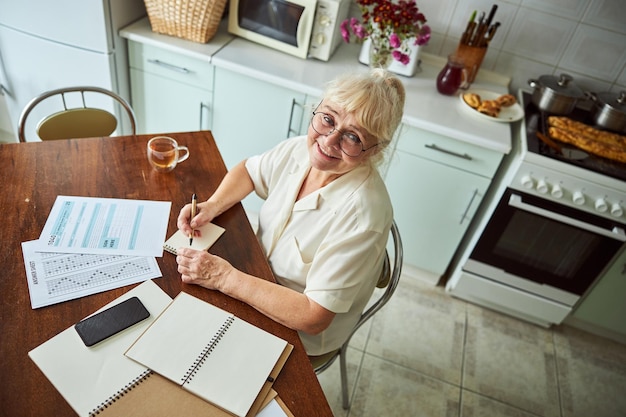 Fröhliche alte Frau, die zu Hause Notizen macht und Papierkram erledigt