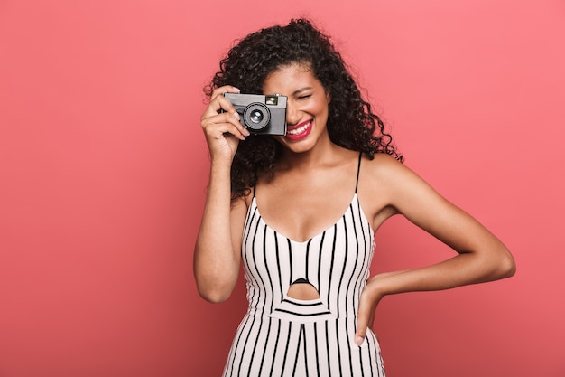 fröhliche amerikanische frau mit lockigem haar, die auf retro-kamera isoliert über rosa wand fotografiert