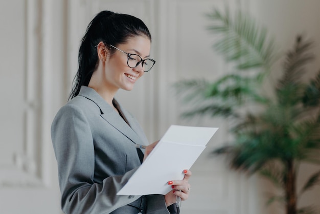 Fröhliche Anwältin schreibt in Unternehmensdokumenten, trägt eine optische Brille, formelle Kleidung, steht im eigenen Schrank, entwickelt neue Strategie, hat einen glücklichen Ausdruck, Menschen, Karriere, Beschäftigungskonzept