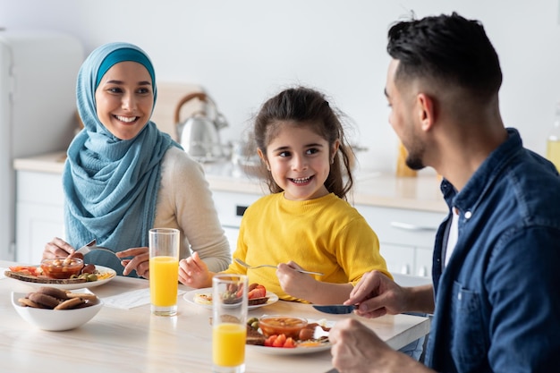 Fröhliche arabische dreiköpfige Familie, die zu Hause leckeres Frühstück in der Küche isst, fröhliche muslimische Eltern und ihre kleine Tochter am Tisch sitzen, leckeres Essen genießen und Orangensaft trinken