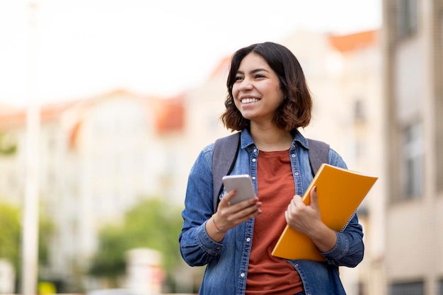 Fröhliche arabische Studentin mit Smartphone und Arbeitsmappen im Freien