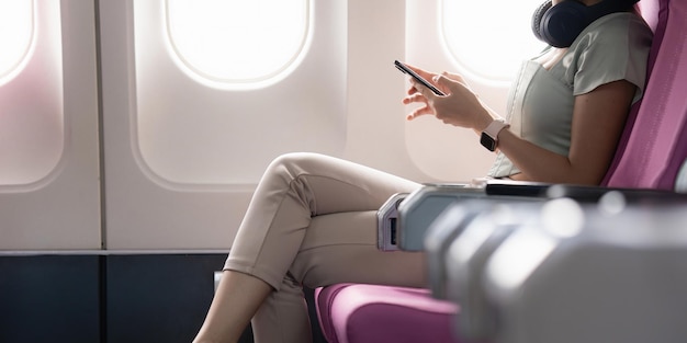 Fröhliche Asiatin sitzt im Flugzeug und benutzt Handy, während sie auf Reisen geht