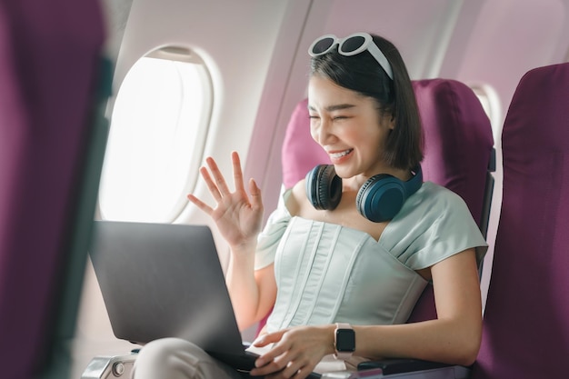 Fröhliche Asiatin sitzt im Flugzeug und benutzt Laptop, während sie auf Reisen geht