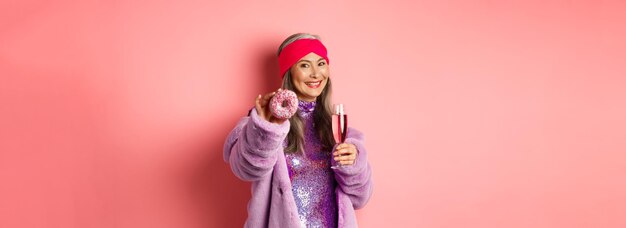 Fröhliche asiatische ältere Frau in schicker Kleidung, die Champagner trinkt und Ihnen Donut gibt, verlängert die Hand mit d