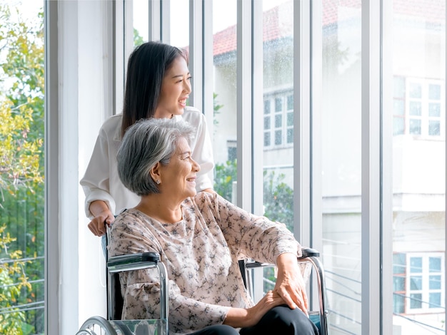 Fröhliche asiatische ältere Frauen, Mutter oder Großeltern im Rollstuhl, die sich um die Pflege kümmern, lächelnde junge weibliche Tochter oder Enkelkind schauen aus dem Fenster und unterstützen zu Hause die Seniorengesundheit