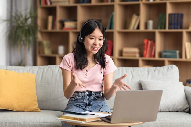 Fröhliche asiatische Dame mit Kopfhörern und Laptop, die online von zu Hause aus lernt und gestikuliert