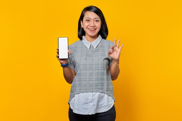 Fröhliche asiatische Frau, die einen leeren Bildschirm des Smartphones zeigt und ein Okay-Zeichen auf gelbem Hintergrund gestikuliert