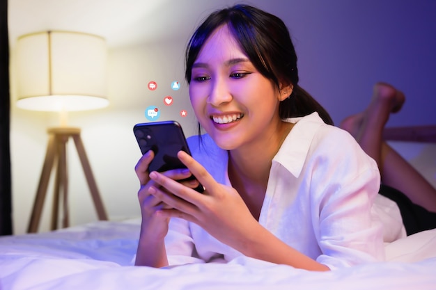 Fröhliche asiatische Frau, die Smartphone mit Benachrichtigungssymbolen von Social Media im Schlafzimmer in der Hausnacht verwendet.