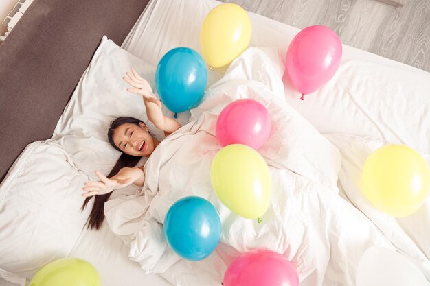 Fröhliche asiatische Frau, die zu Hause mit bunten Luftballons im Bett liegt