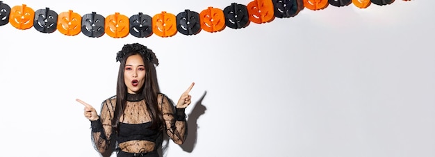 Foto fröhliche asiatische frau im gotischen spitzenkleid, die halloween feiert, mit den fingern zur seite zeigt und vor weißem hintergrund mit kürbisparty-luftschlangen steht