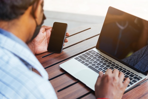 Fröhliche asiatische handschwarze Person, die mit Laptop-Computer arbeitet und Nachrichten von moderner Smartphone-Gerätetechnologie mit leerem Bildschirm liest und Online-Netzwerke im Café-Café-Shop verbindet