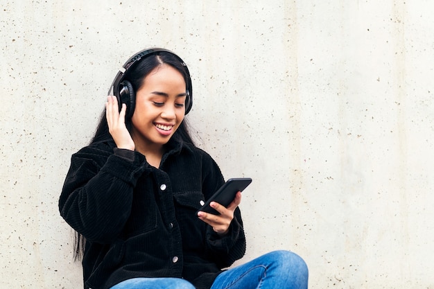 Fröhliche asiatische junge Frau mit Kopfhörern, die Musik vom Telefon hört, das draußen auf ihrem Skateboard sitzt und sich an eine Betonwand lehnt