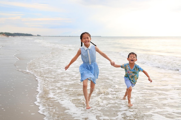 Fröhliche asiatische junge Schwester und kleiner Bruder, die Spaß daran haben, bei Sonnenaufgang am tropischen Sandstrand zusammenzulaufen Fröhlicher Familienjunge und -mädchen genießen die Sommerferien