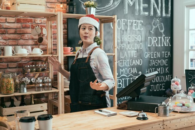 fröhliche asiatische koreanerin in roter weihnachtsmütze, die weihnachtsfeiertage in der bartheke feiert. sorglose Kellnerin in niedlichem Dekor auf dem Kopf, die während der Arbeit im Café tanzt und Spaß hat.