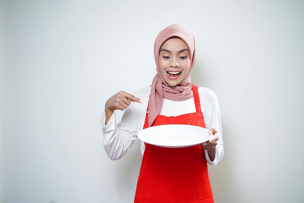 Fröhliche asiatische muslimische Frau in roter Schürze, die auf einen leeren Teller zeigt Lebensmittelwerbung Kochkonzept